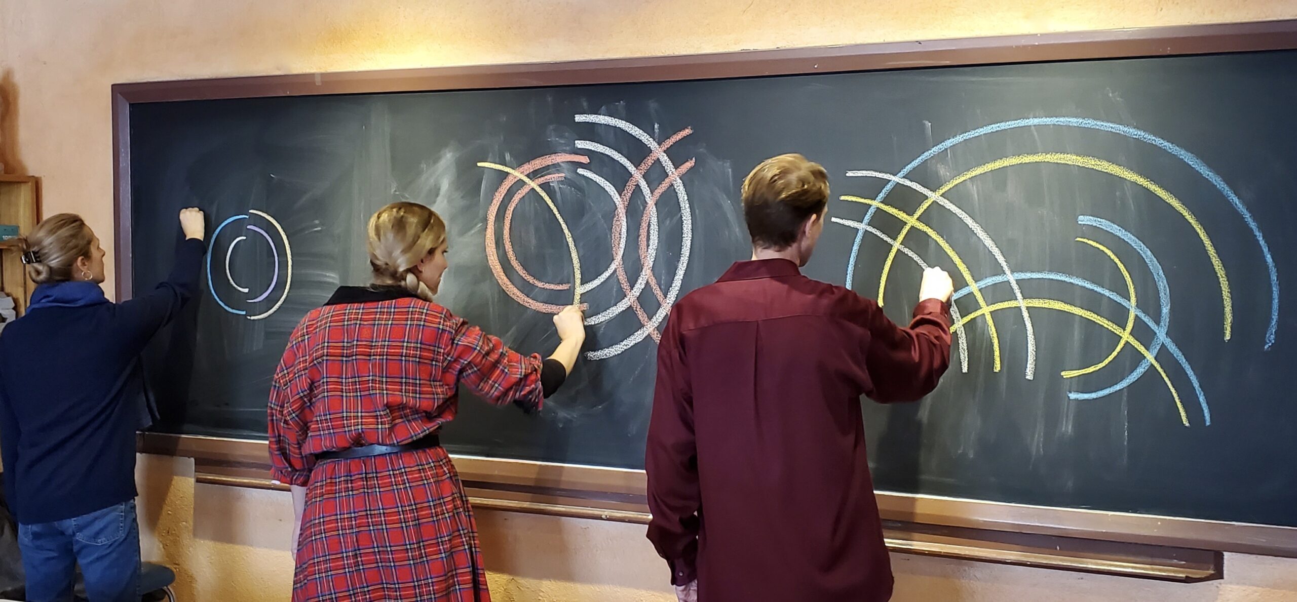 Two female students on the left and one male student on the right have their backs towards the camera as they draw bright curved lines on a chalkboard.