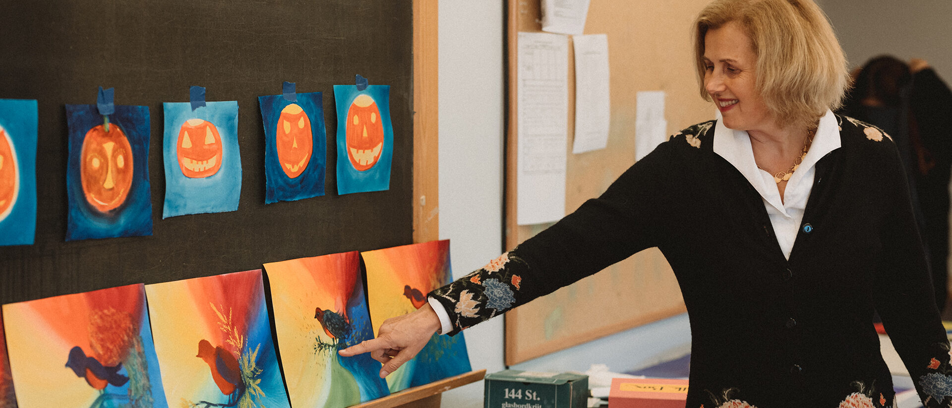 Painting instructor Martina Muller standing at chalkboard where student work is displayed pointing to one of the colorful paintings of a bird