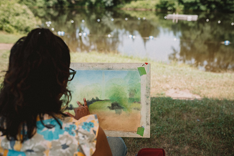 Back of woman smudging oil pastel piece of a pond in front of her.