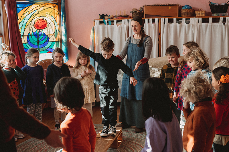 Kindergarten class circling a student walking across a wooden beam as teacher helps guide him.