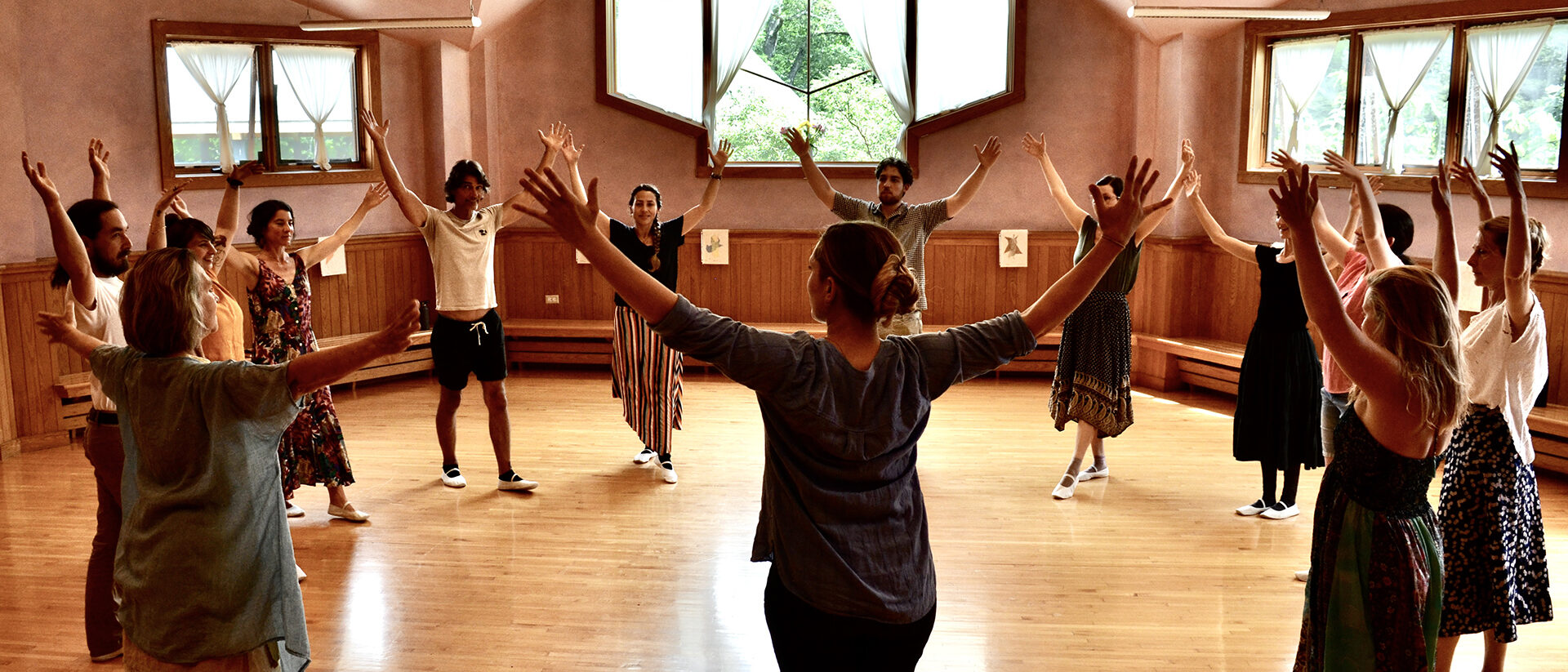 students gathered in a circle in a sunlit room practicing eurythmy; every student has their hands raised over their head